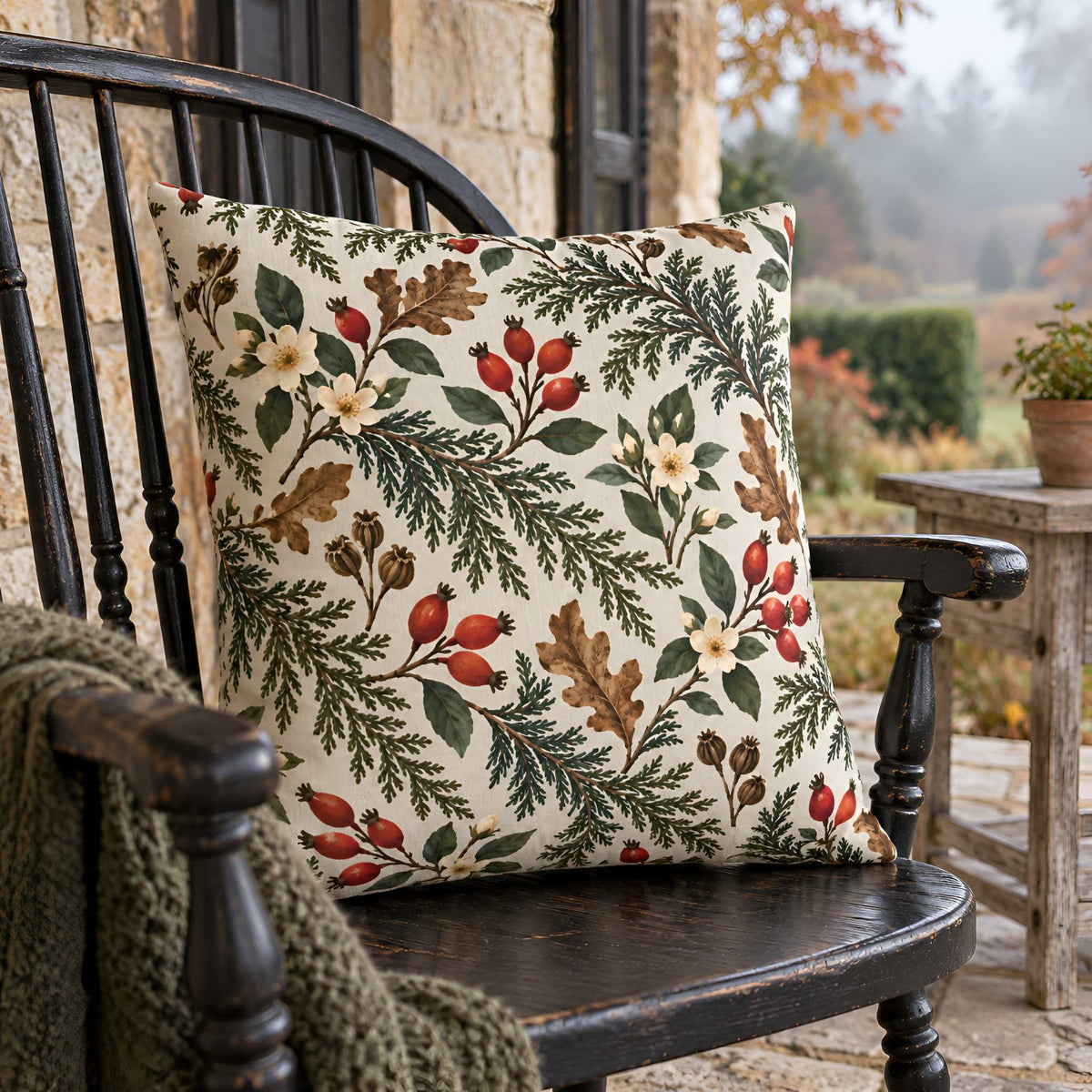 A square decorative throw pillow with a detailed botanical pattern of pine branches, oak leaves, and red berries on a cream background, resting on a dark wooden rocking chair in an outdoor patio setting.