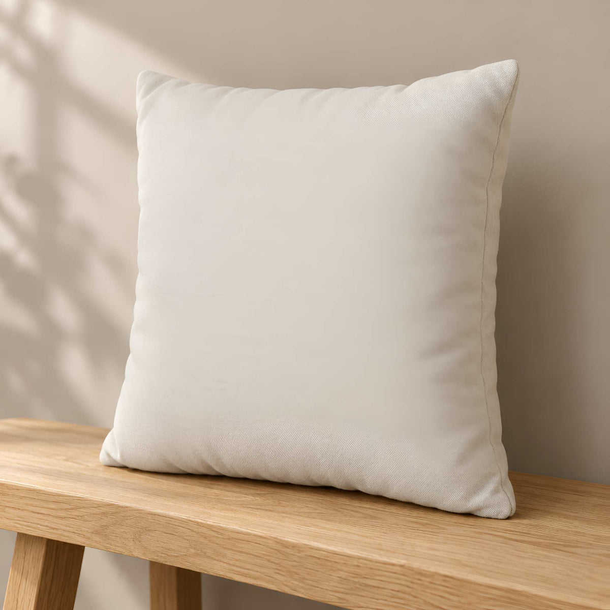A square throw pillow cover resting on a light wood bench against a neutral beige wall with soft leaf shadows.