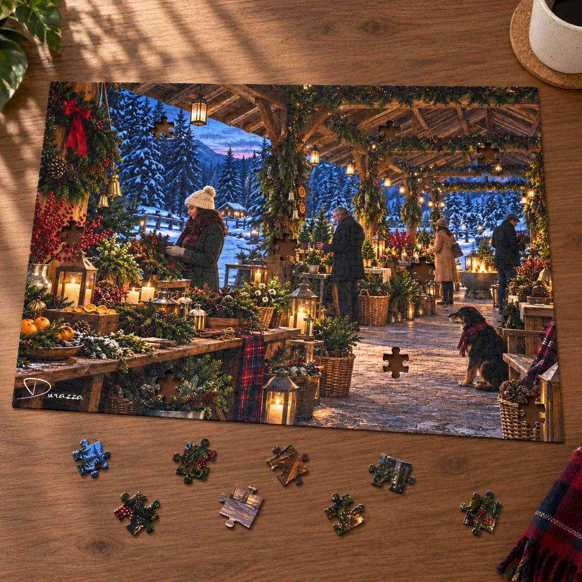 A completed rectangular jigsaw puzzle showing a festive Christmas market scene with people, lanterns, garlands, and trees, placed on a wooden tabletop. Several loose puzzle pieces are scattered around it, and part of a plant and a cup are visible at the edges.